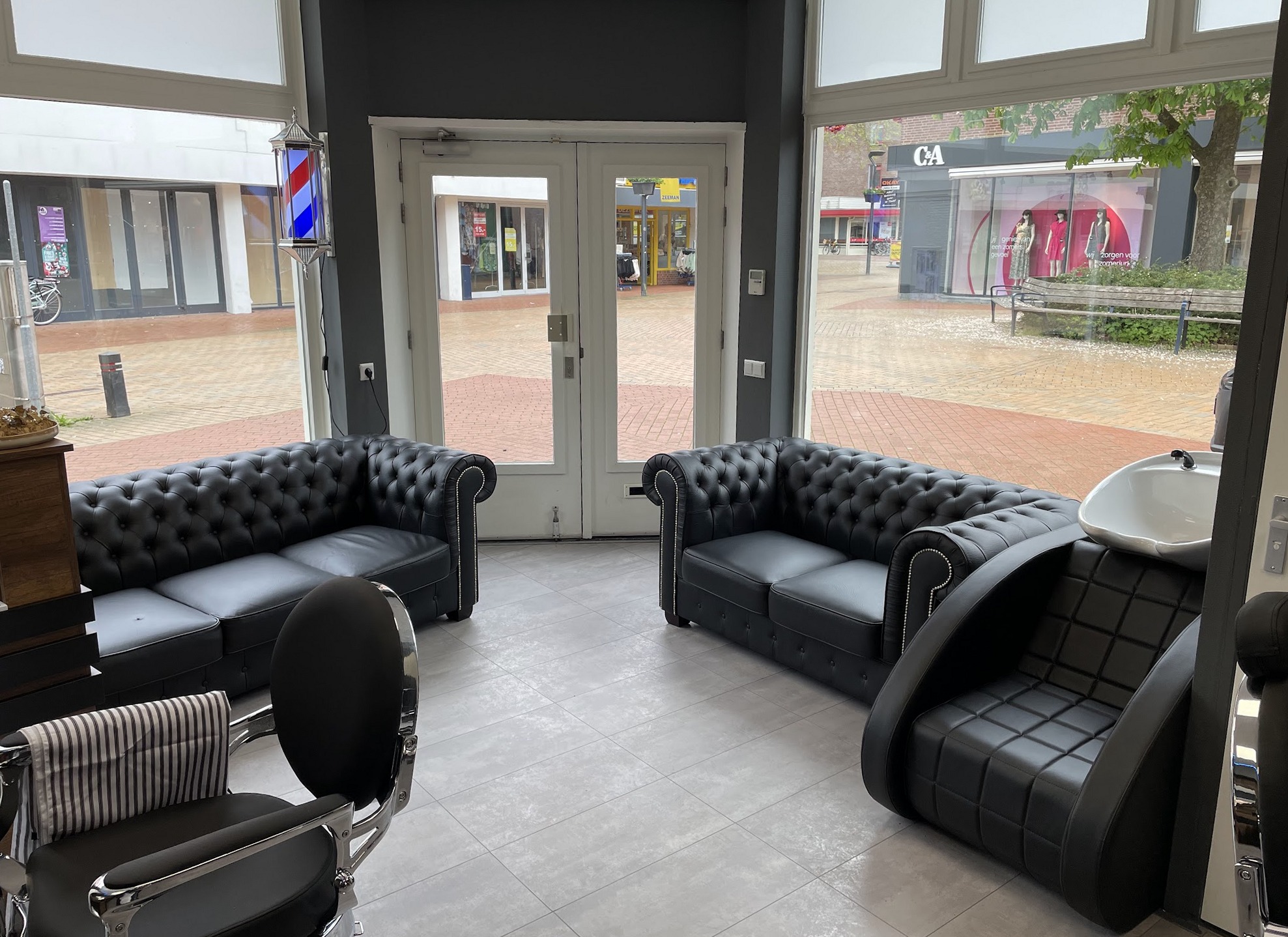 Wachtruimte met zwarte Chesterfield banken, een kappersstoel en een wasbak, met door de glazen deuren de Noorderbuurt en een kapperspaal